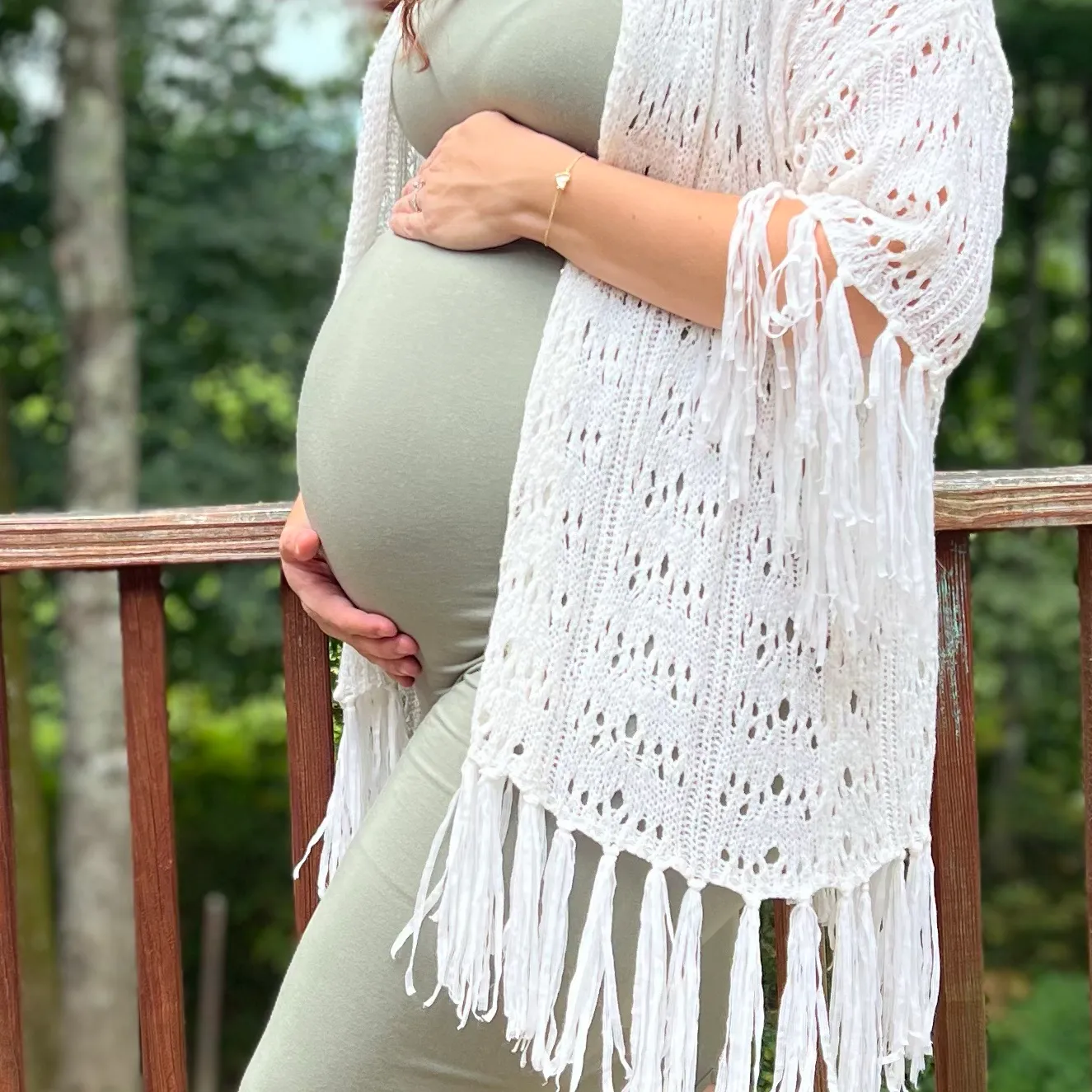 The gardener's hands resting on her bump, in a sage green dress and a white crochet shawl, with trees and a wooden railing behind her.